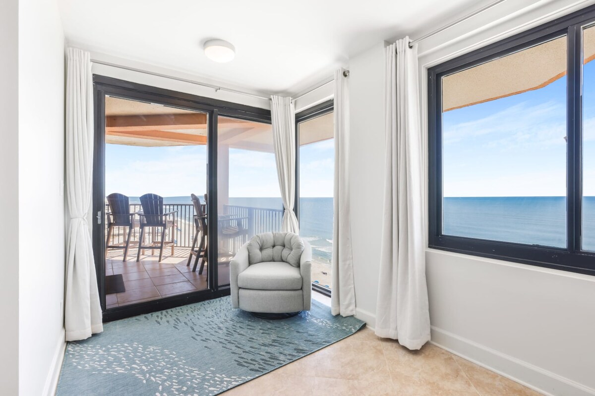 The image features a comfortable seating area with a soft armchair, positioned near expansive windows that frame unobstructed beach views. Sheer curtains gently drape the windows, allowing natural light to fill the space. A textured area rug complements the neutral-toned tiled floor.