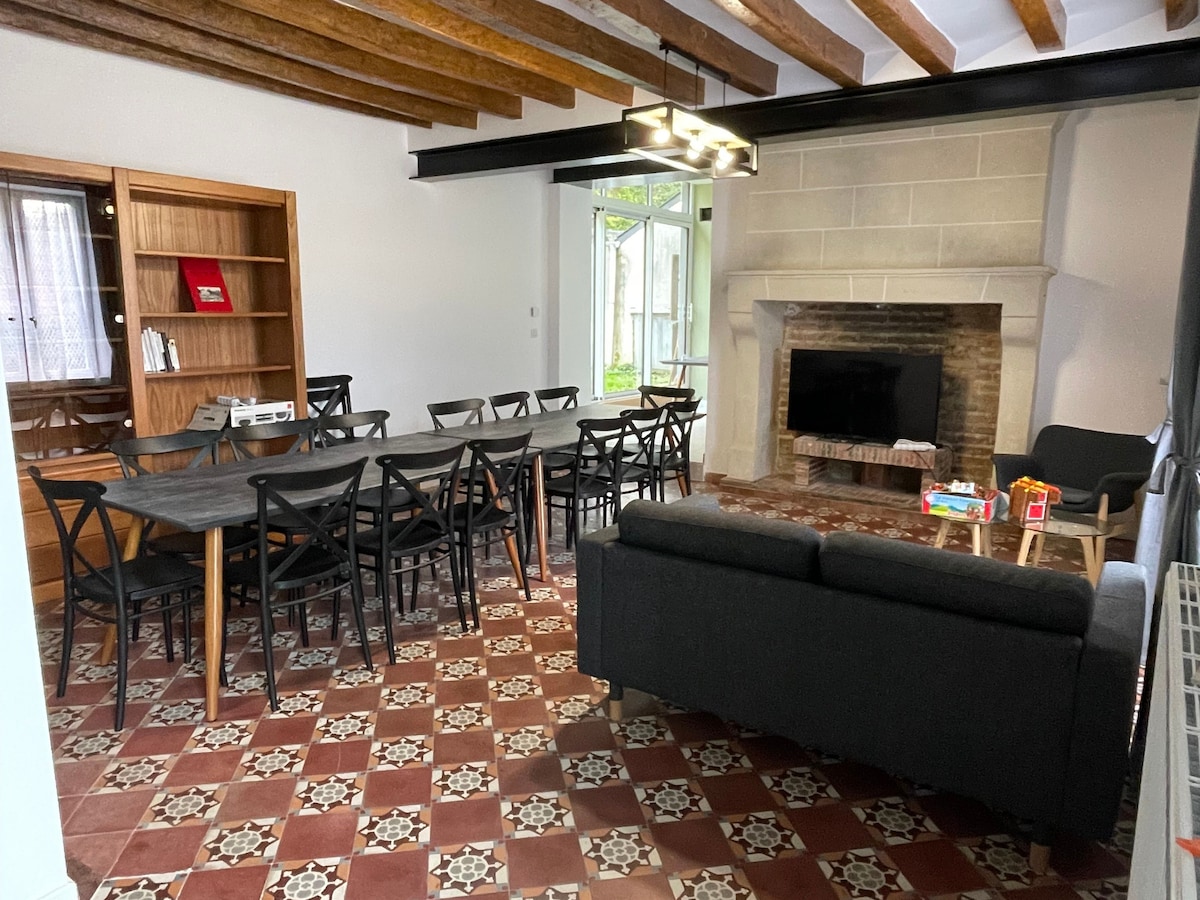 A spacious living room features a long dining table surrounded by black chairs. A comfortable sofa is positioned nearby. A television sits on a stone fireplace, with large windows allowing natural light to fill the room. Decorative tiled flooring adds character.