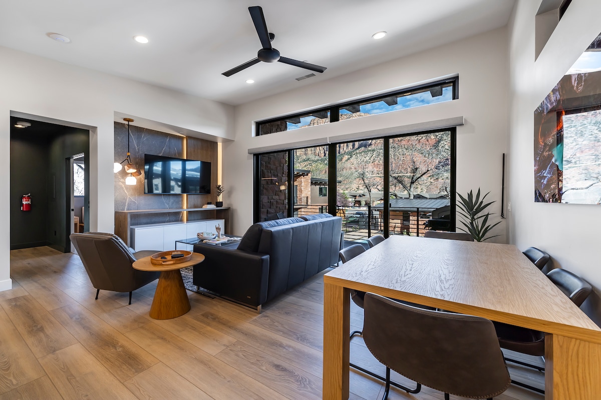 The living area features a sectional sofa facing a wall-mounted television. A round wooden coffee table sits in front, and a modern dining table with chairs is positioned nearby. Large windows provide natural light and showcase views of the surrounding landscape.