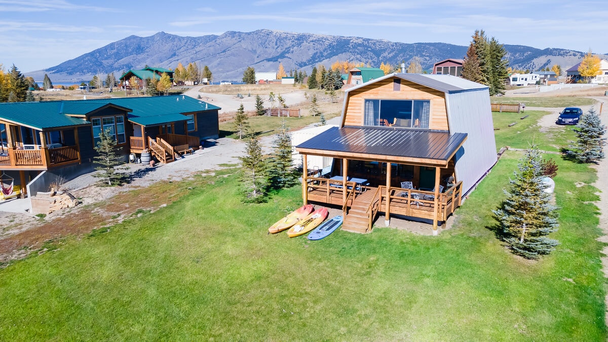 An aerial view showcases a modern cabin with a spacious deck, surrounded by green grass and a mountainous backdrop. Kayaks and paddle boards are arranged neatly near the entrance, complementing the outdoor space. Additional cabins are visible in the distance, highlighting a serene setting.