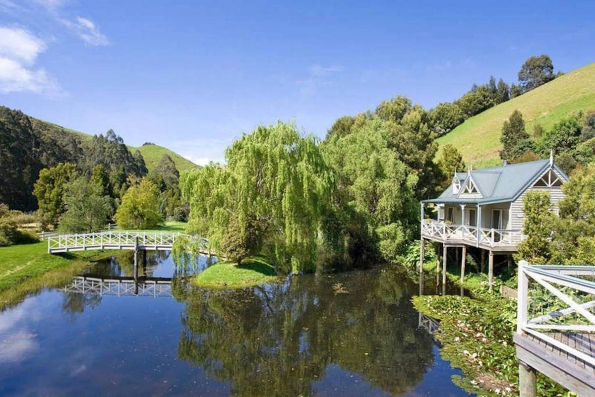 The exterior of Little Plum Cottage is depicted alongside a tranquil ornamental lake. A wooden bridge crosses over the water, surrounded by lush greenery and hills in the background, creating an inviting and peaceful landscape.
