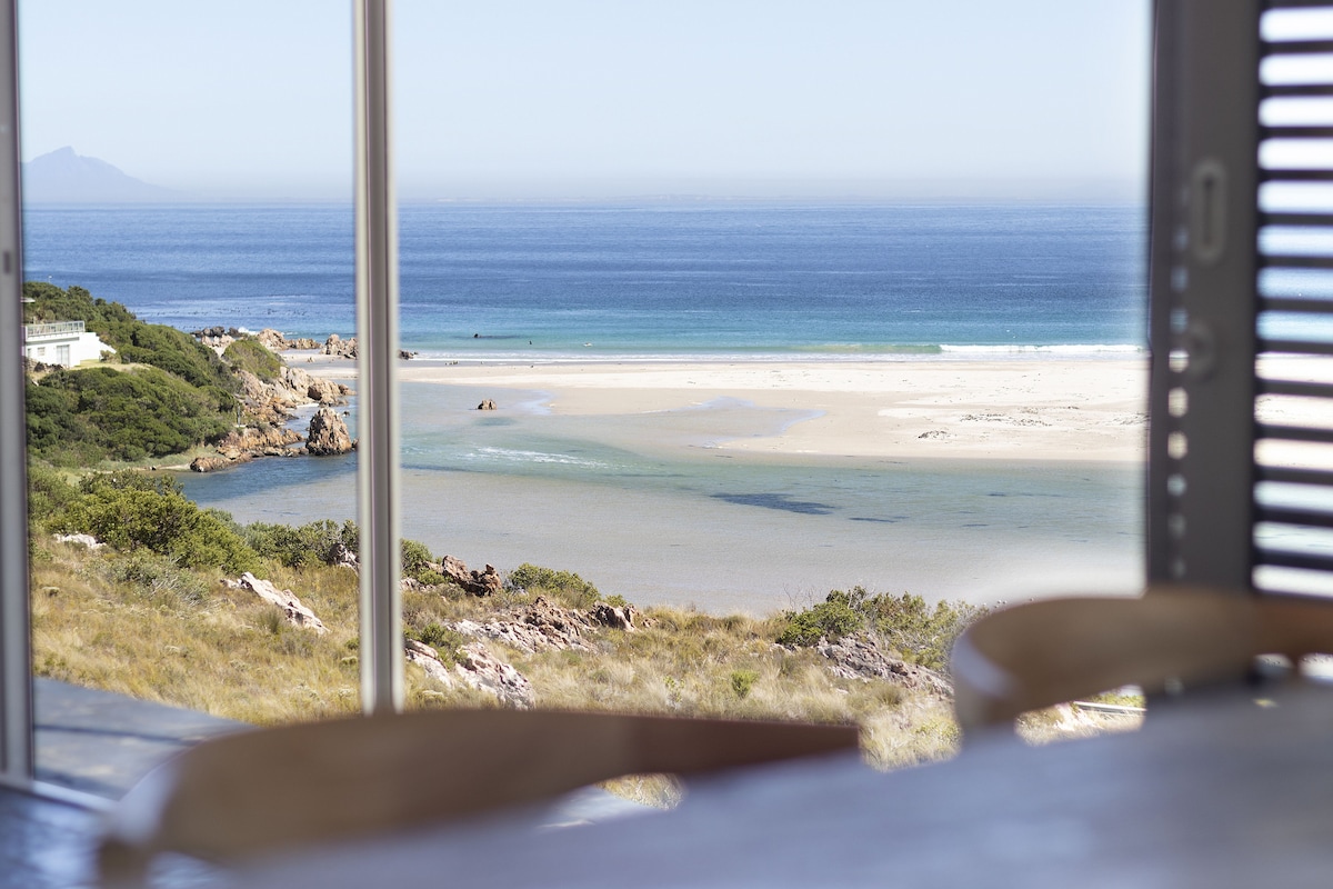 A serene view of the coastline is framed by large glass windows. Soft waves gently lap against the sandy shore, while the vibrant blue hues of the ocean and sky create a calming atmosphere. Nearby greenery adds to the natural beauty of the setting.