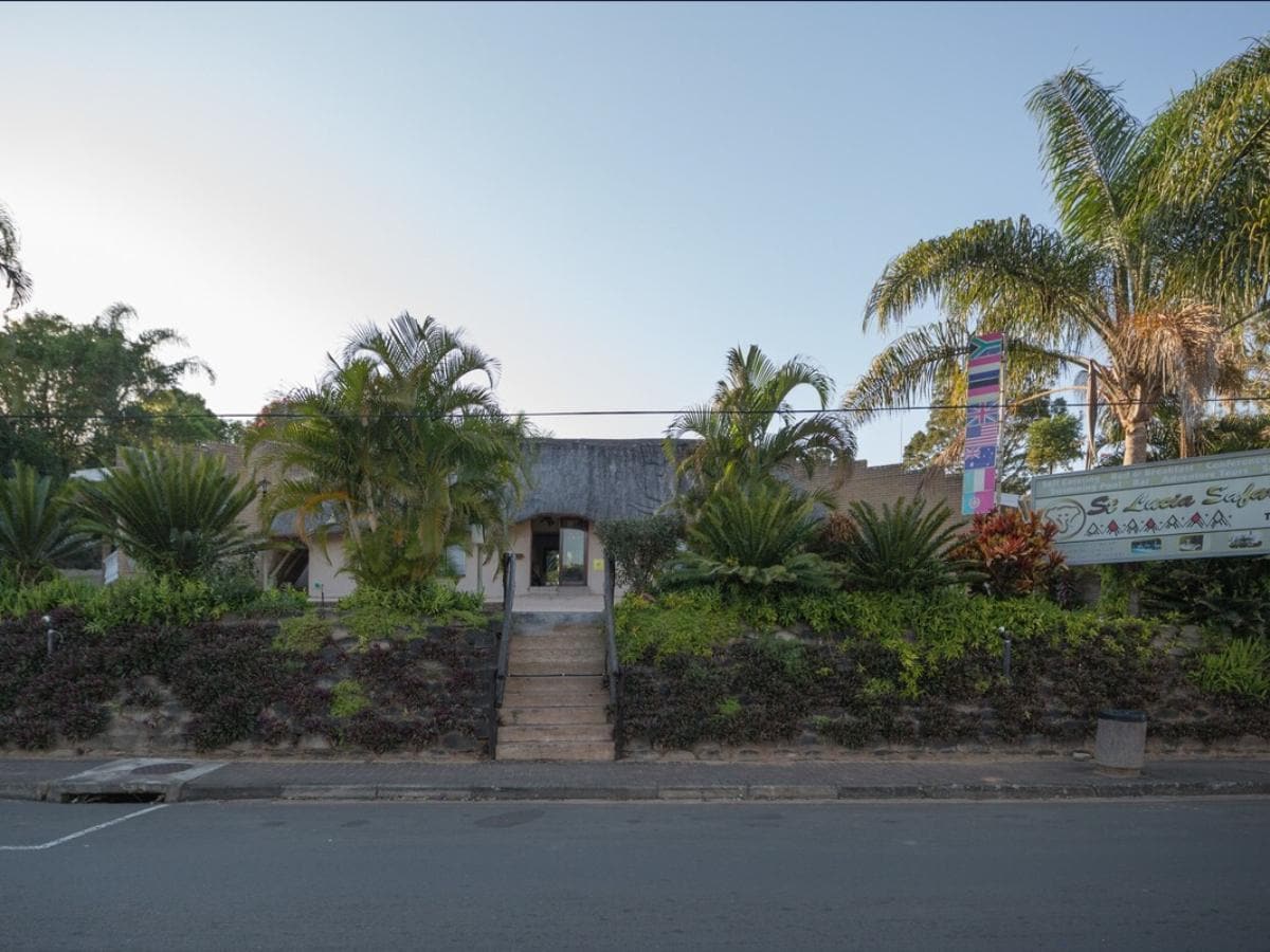 A low-rise building features a thatched roof, surrounded by lush greenery and palm trees. A stone staircase leads to the entrance, which is framed by colorful banners and tropical plants, enhancing the welcoming facade.