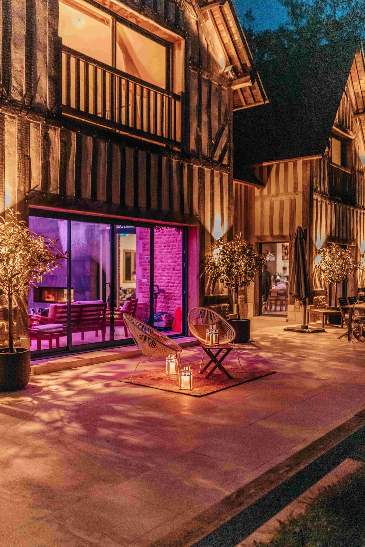 An inviting outdoor space is illuminated by soft lighting, featuring two chairs positioned on a patterned rug. The patio area is bordered by lush greenery, while large glass doors provide a glimpse into the interior living space, reflecting a warm ambiance.