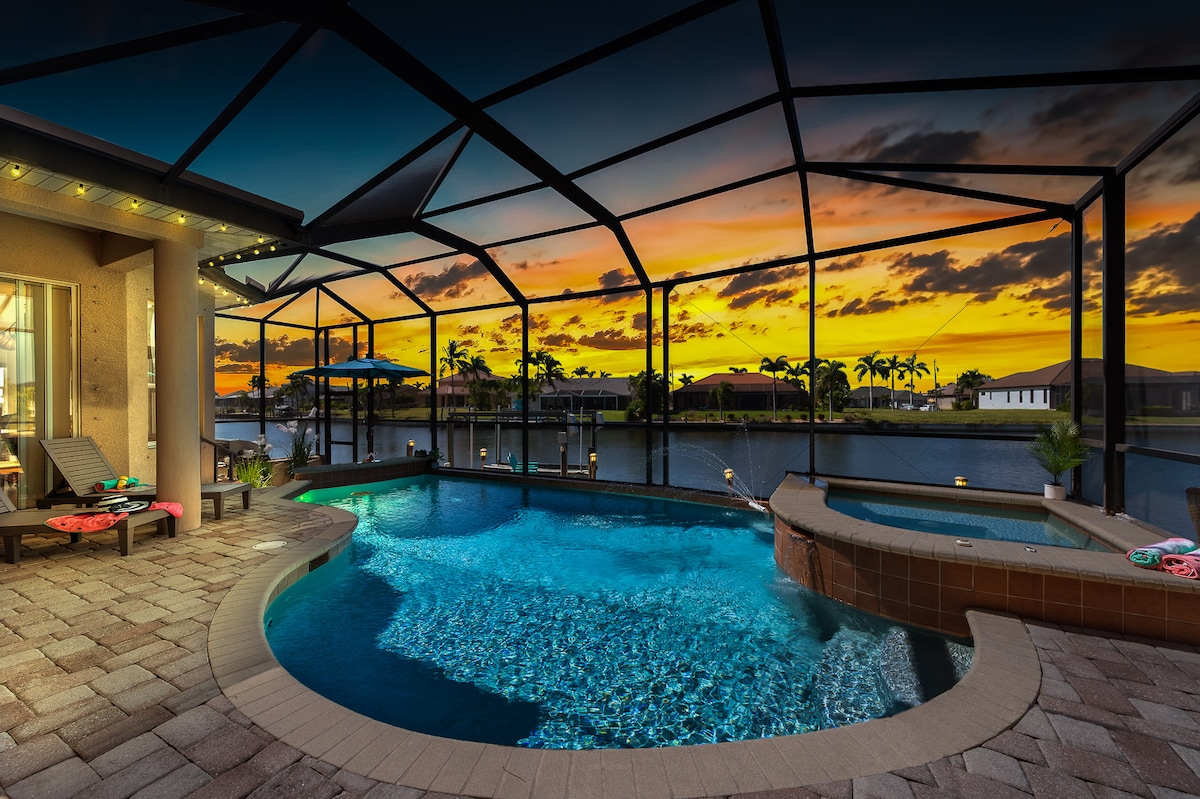 An inviting outdoor space features a refreshing pool and spa surrounded by a screened enclosure. The vibrant sunset casts a colorful glow over the water, enhancing the serene canal view. Comfortable seating is arranged nearby, perfect for relaxation and enjoying the tranquil atmosphere.