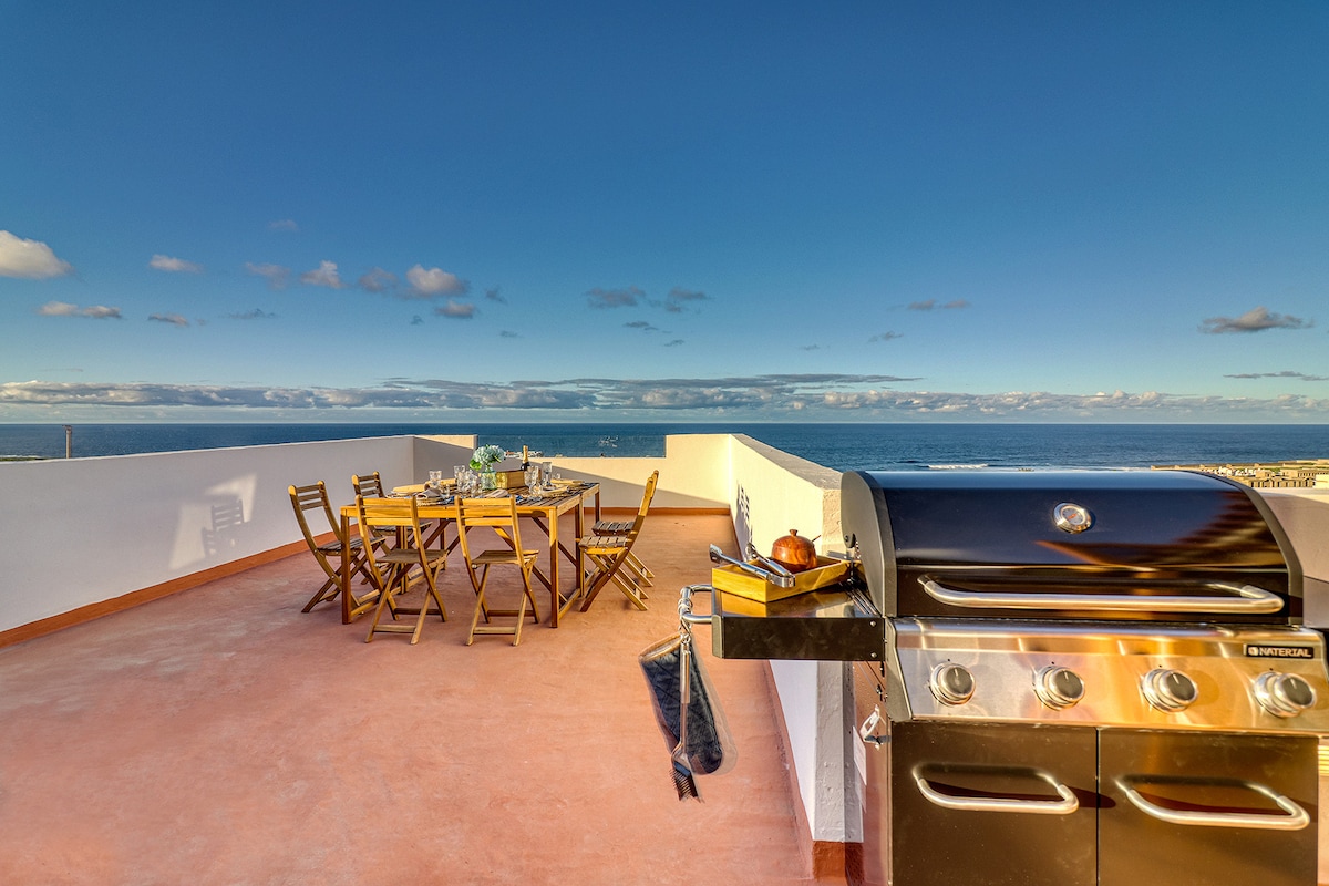 A spacious rooftop terrace features a large dining table surrounded by wooden chairs, offering views of the ocean. A gas grill is positioned nearby, ideal for outdoor cooking. The area is bathed in natural light, with a clear blue sky overhead.
