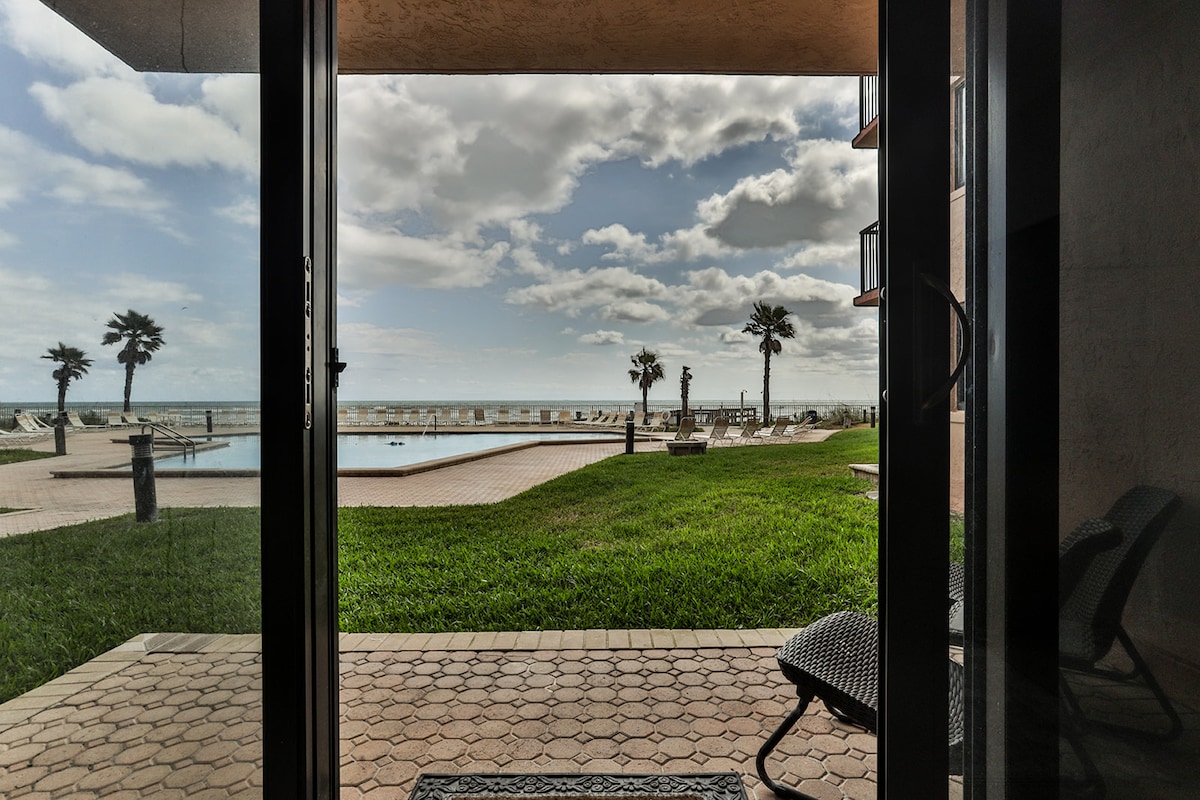 A view from the ground floor showcases a spacious outdoor area leading to the ocean. Palm trees frame the scene, while a tranquil pool is visible in the foreground. The expansive coastline stretches across the horizon under a partly cloudy sky.