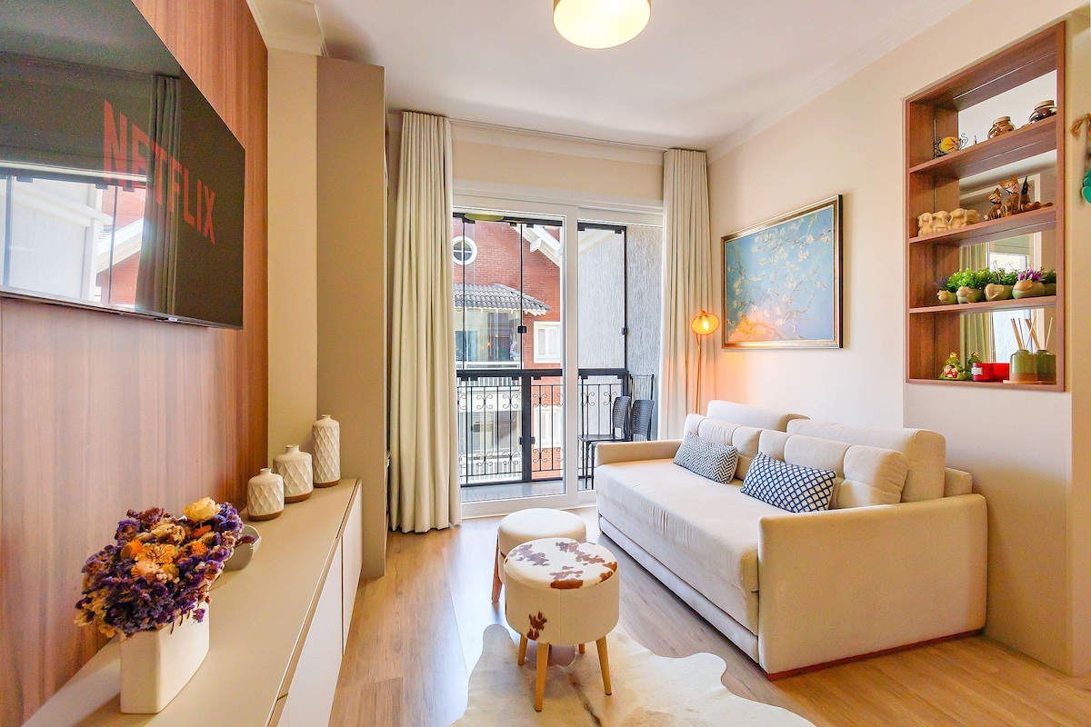 A light-filled living area showcases a comfortable sofa with decorative cushions, positioned in front of a wall-mounted Smart TV. Large glass doors lead to a balcony, enhancing the space with natural light. A small decorative table and vibrant artwork add visual interest.