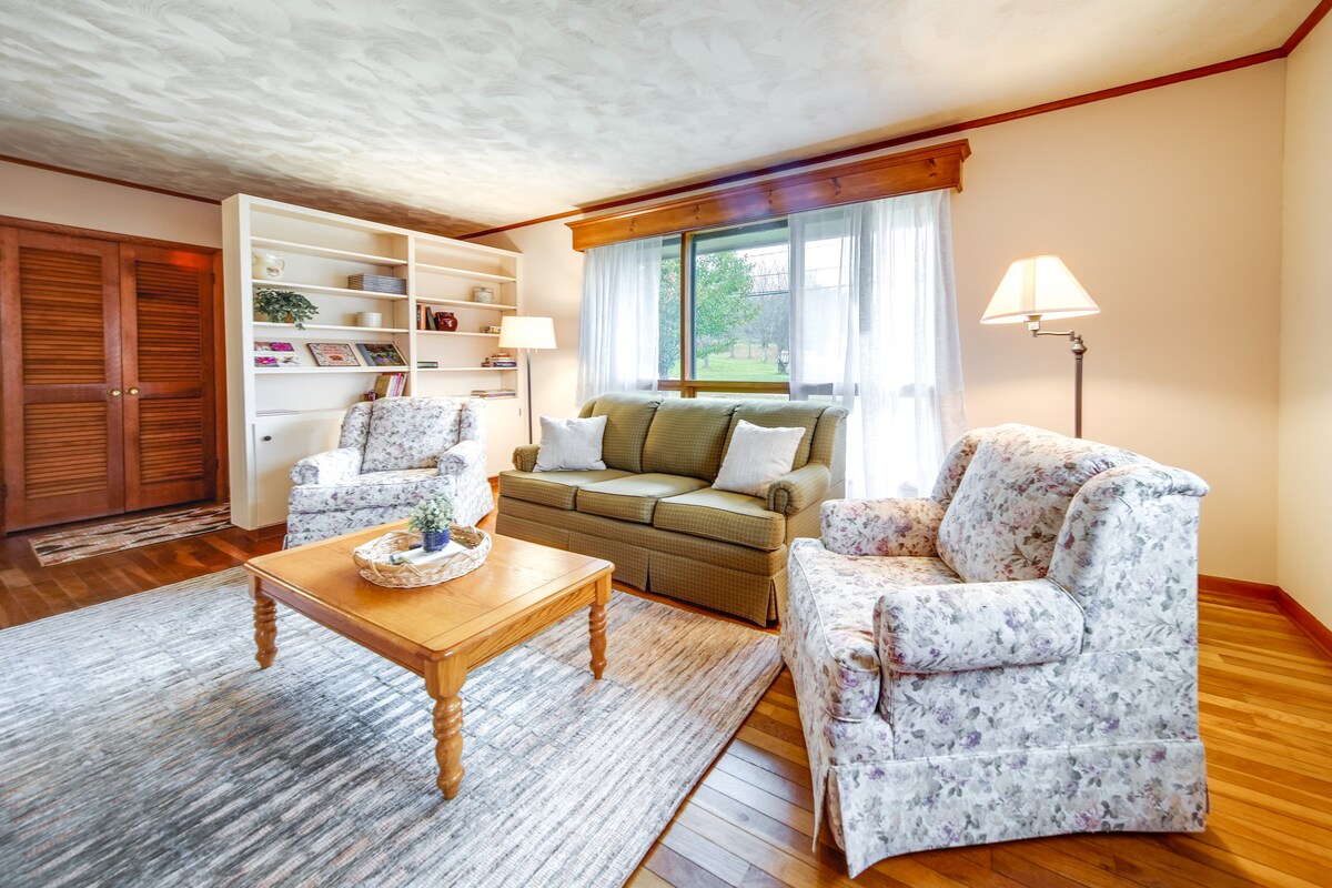 A comfortable living area features a cozy green sofa and two floral-patterned armchairs. A wooden coffee table is centered on a soft area rug. Natural light filters through a large window, complemented by light curtains, while built-in shelves showcase decorative items.