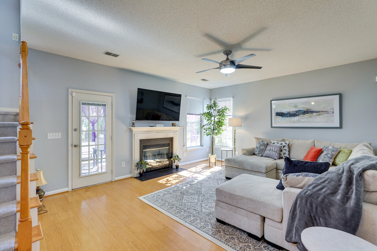 A spacious living area features a large sectional sofa, complemented by a cozy blanket and assorted pillows. A ceiling fan contributes to air circulation. Natural light enters through open patio doors, illuminating a fireplace beneath a mounted television. Neutral tones create a calm ambiance.