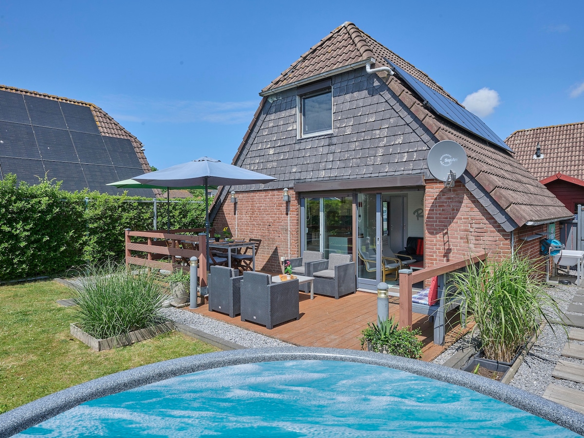 A modern house with a sloped roof is surrounded by a landscaped garden. A cozy terrace with outdoor seating and an umbrella overlooks a heated hot tub, creating a relaxing outdoor space. Lush greenery and simple décor enhance the inviting atmosphere.