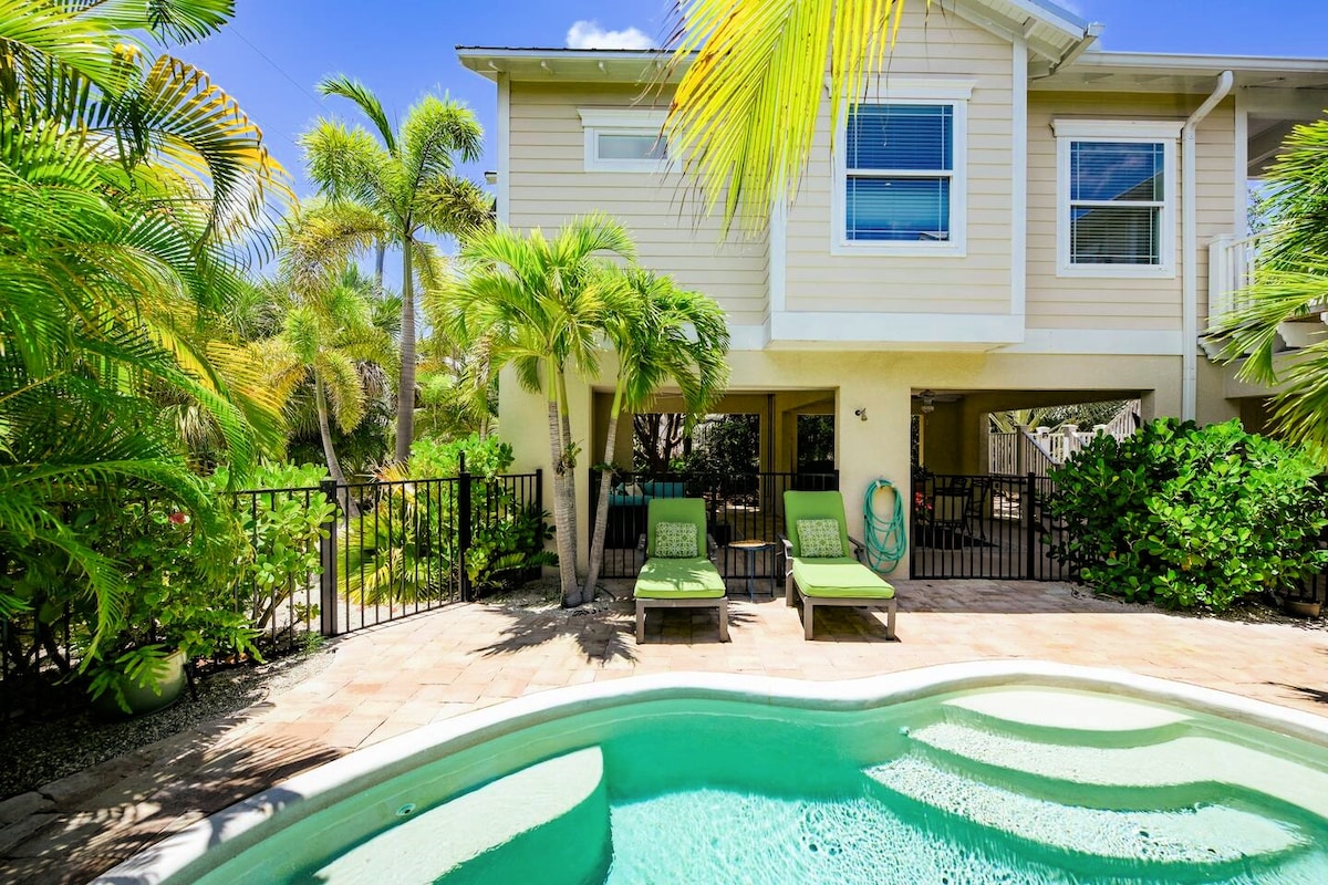 A private yard features a heated pool surrounded by lush tropical landscaping. Two lounge chairs sit by the water, inviting relaxation under the warm sun. The elevated home is visible above, offering a comfortable living space filled with natural light.