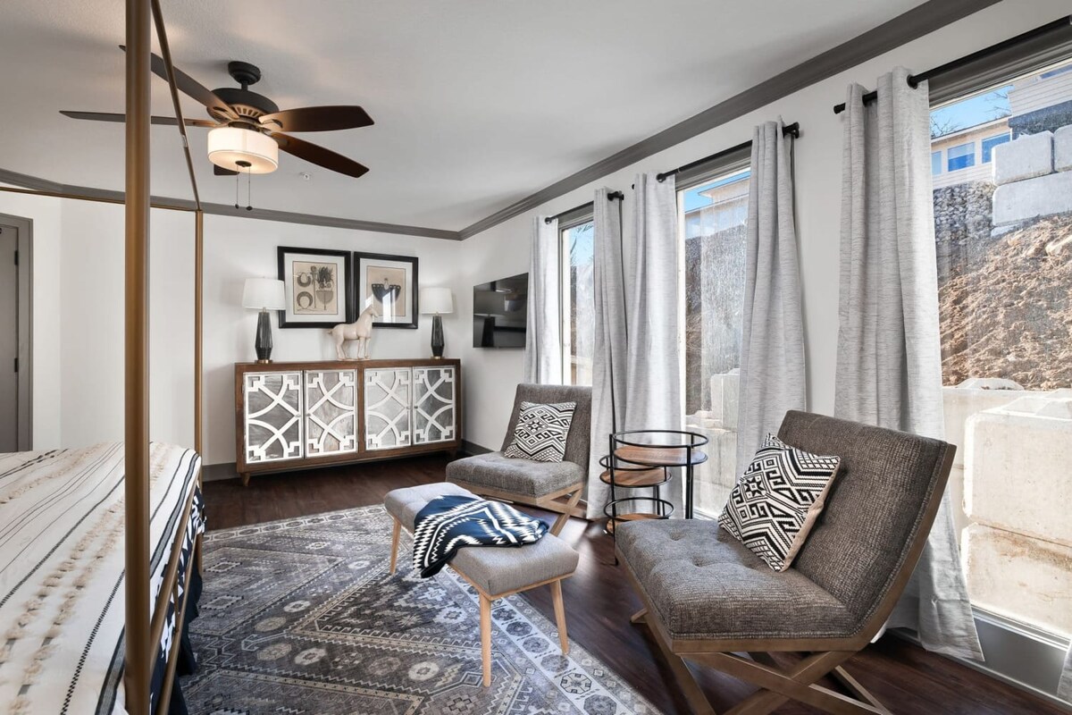 A cozy seating area is highlighted by two textured armchairs with geometric patterns. A rug with intricate designs anchors the space, while a stylish media console complements the room's decor. Light filters through expansive windows, providing a warm atmosphere.
