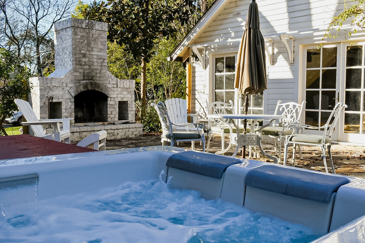 An outdoor seating area features a dining table with an umbrella, surrounded by four chairs. A stone outdoor fireplace stands nearby, contributing to the cozy ambiance. In the foreground, a hot tub bubbles, inviting relaxation in a serene setting.