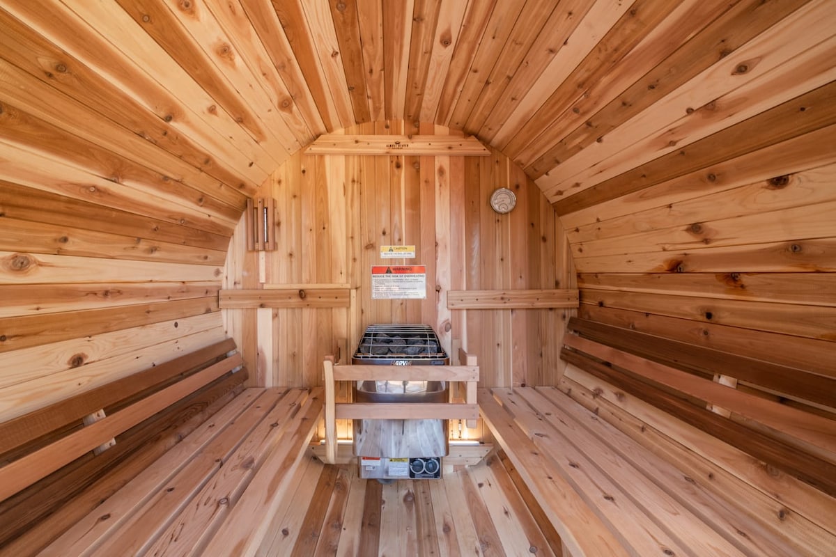 A wooden barrel sauna features warm, natural cedar paneling along the walls and ceiling. A bench runs along the interior, offering ample space for relaxation. A small stove is positioned at the center, with a clock mounted on the wall for timekeeping.