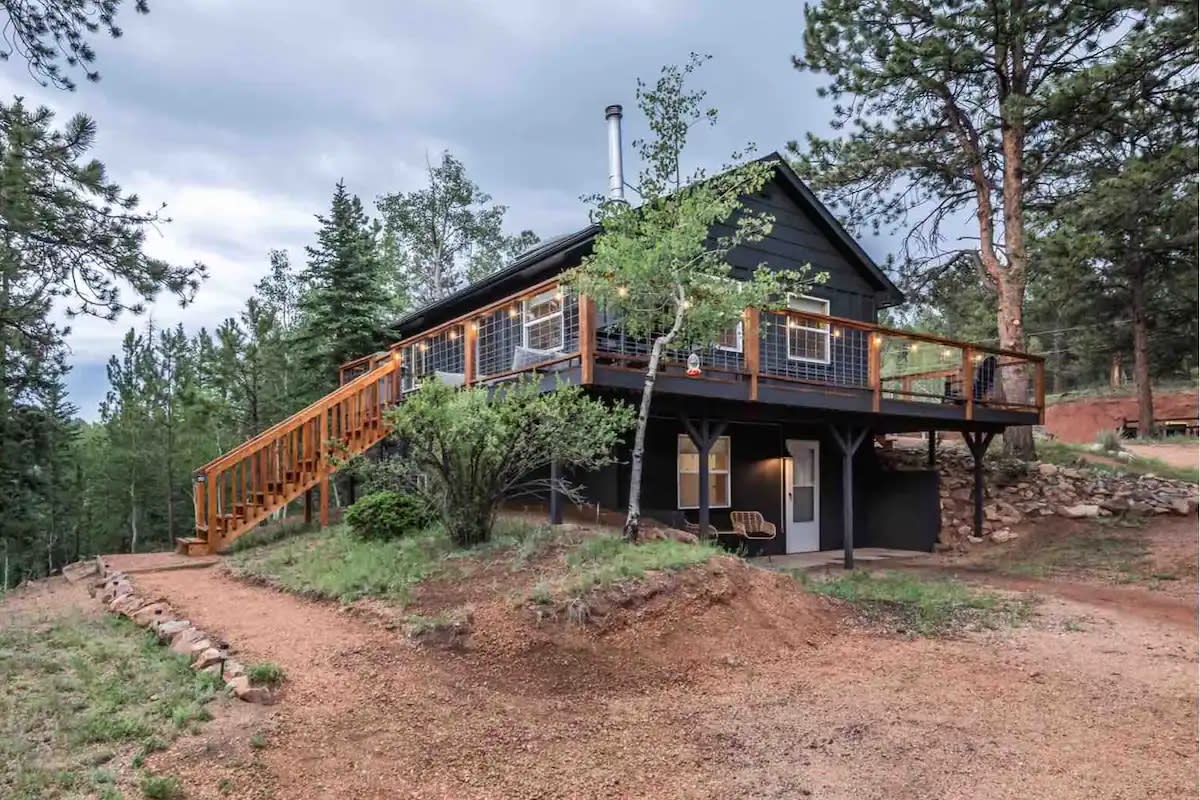 A welcoming cabin is nestled among trees, showcasing a large wrap-around deck and a wooden staircase leading to the entrance. Surrounding greenery and a natural path create an inviting outdoor setting.