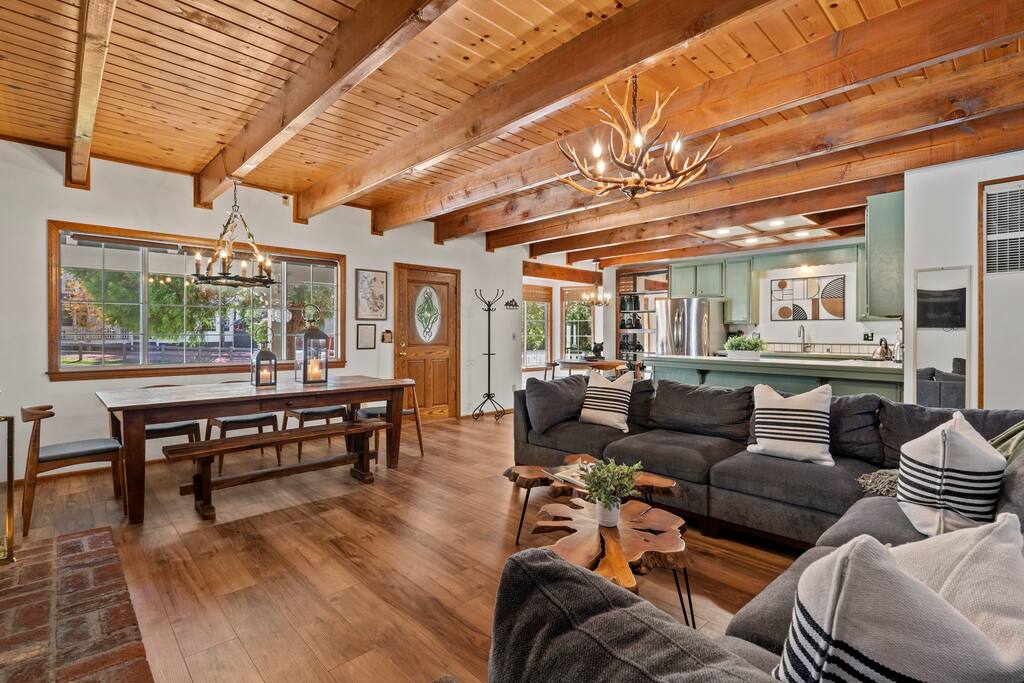 A cozy living space features wood beams and a combination of hardwood floors and stone accents. A large sectional sofa is positioned around a rustic wooden coffee table, complemented by a dining area with a long table and stylish chairs. Abundant natural light enters through large windows.
