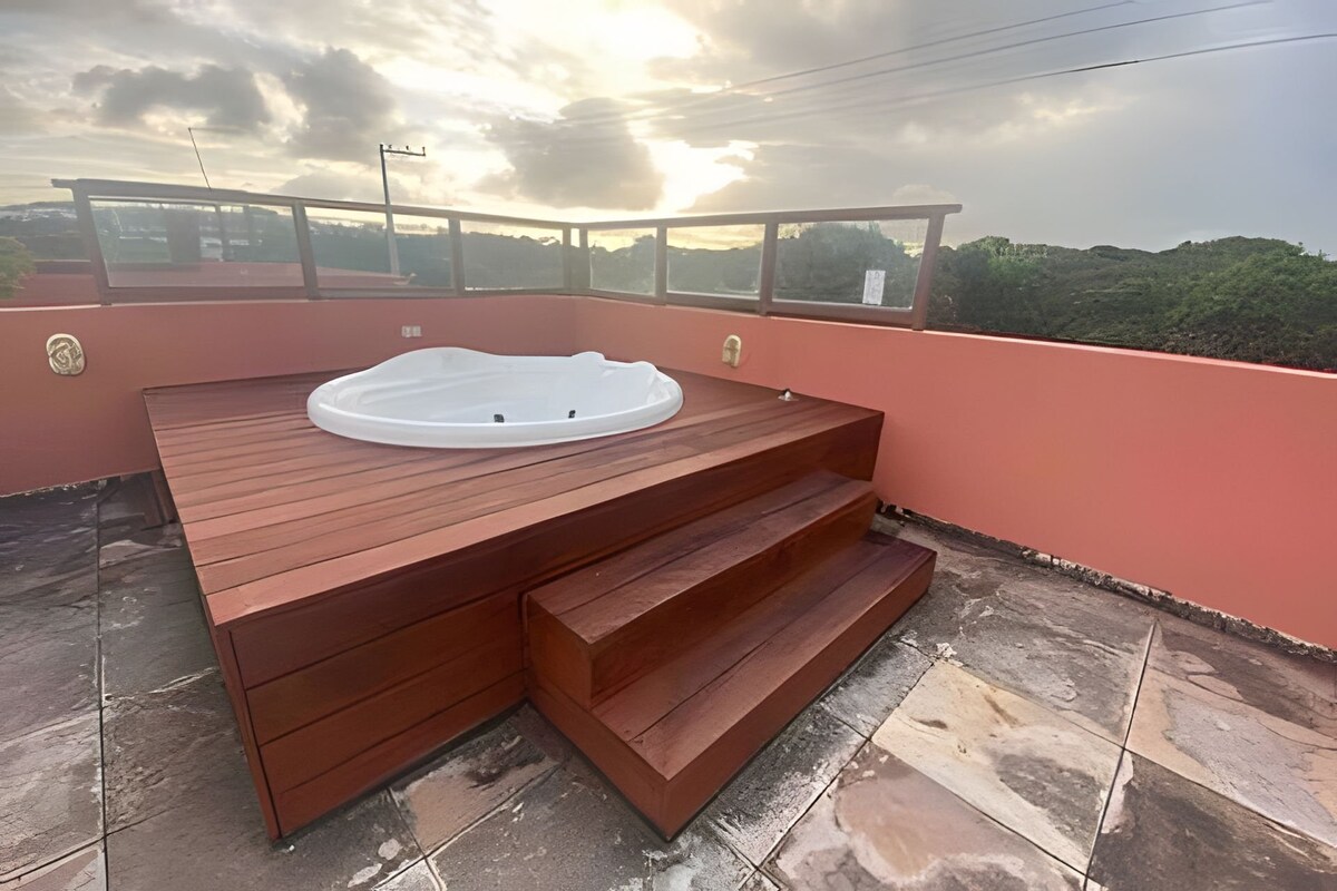A wooden deck area displays a round hot tub, elevated on a platform. The surroundings feature a serene view of nature, with gentle earthy tones in the paneling, complementing the peaceful setting. Steps lead down from the deck, further inviting relaxation.