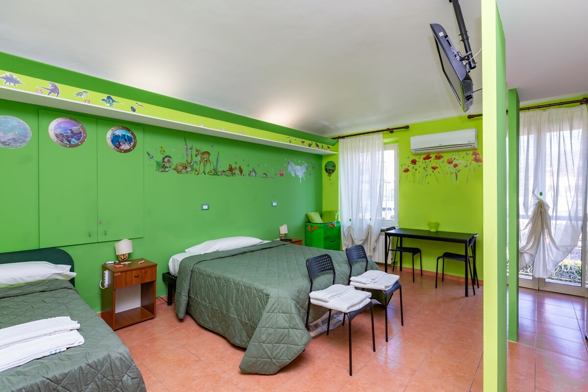 A spacious room features vibrant green walls adorned with nature-themed artwork. Two beds, covered with matching quilted bedding, are positioned alongside a nightstand. A desk and chairs are visible, along with large windows dressed with sheer curtains allowing natural light to illuminate the space.