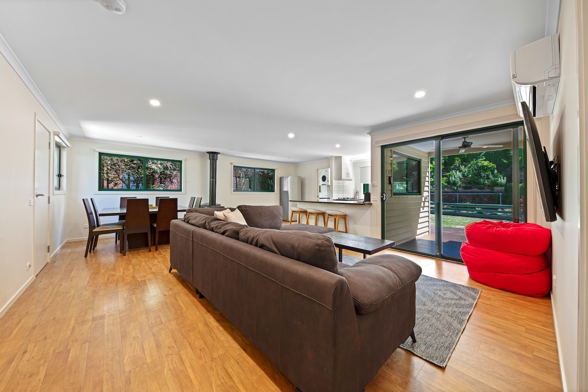 An open-plan living area is visible, featuring a comfortable sofa and a large dining table. Floor-to-ceiling glass doors provide access to an outdoor space, allowing natural light to fill the room. A Smart TV is mounted on the wall, and a fireplace is located in the corner.