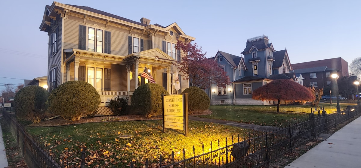 The exterior of The Conkling House is captured at dusk, highlighting its elegant Italianate architecture. The well-maintained lawn is framed by manicured bushes and a classic wrought iron fence. Soft lighting illuminates the entrance, while the welcoming sign stands prominently in the front yard.