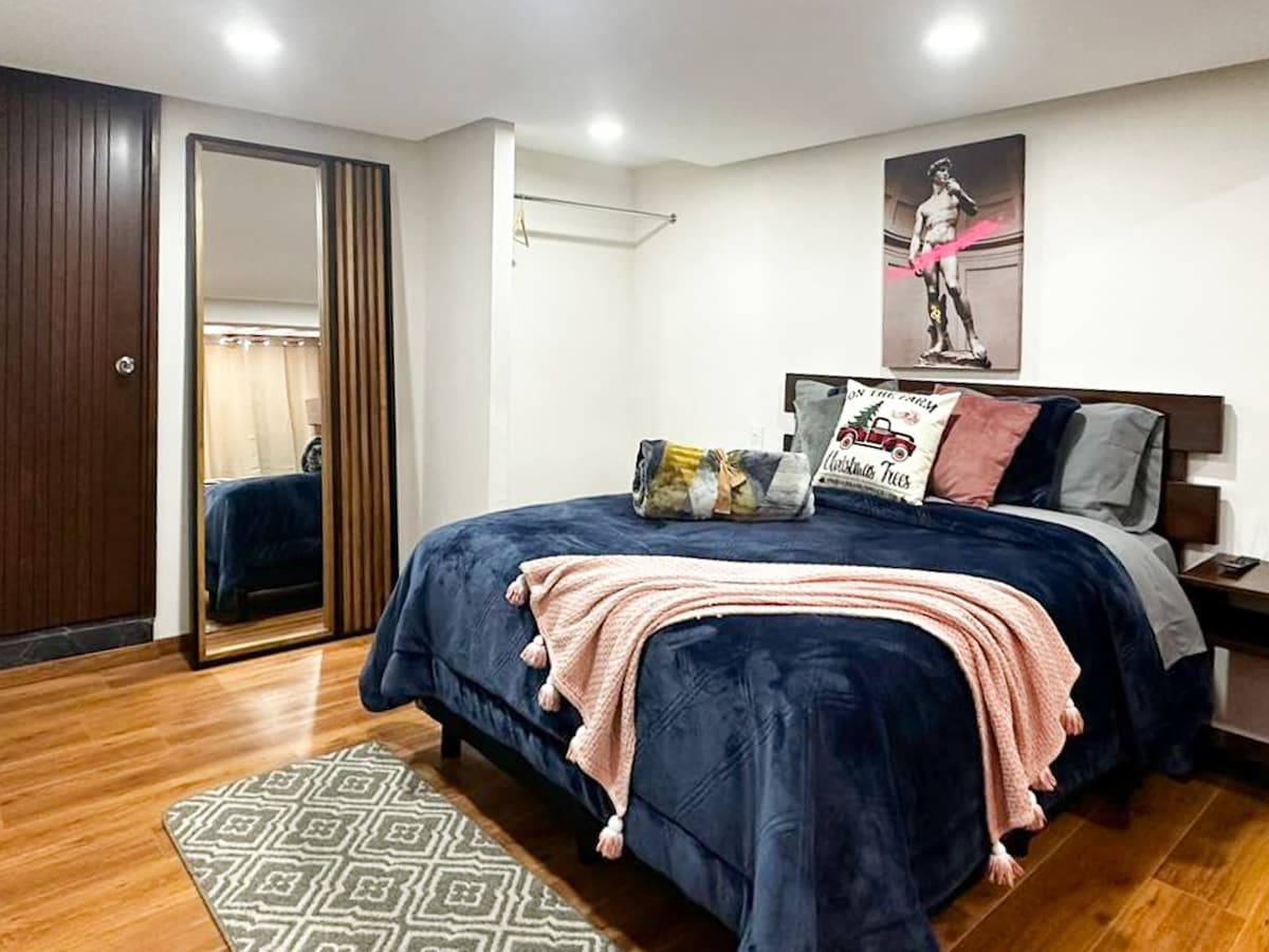 A spacious bedroom is featured, showcasing a large bed draped in navy bedding, accented with decorative pillows. Natural wood flooring complements the room's neutral tones. A full-length mirror and a closet provide practical storage, while a colorful artwork adorns the wall, adding character.