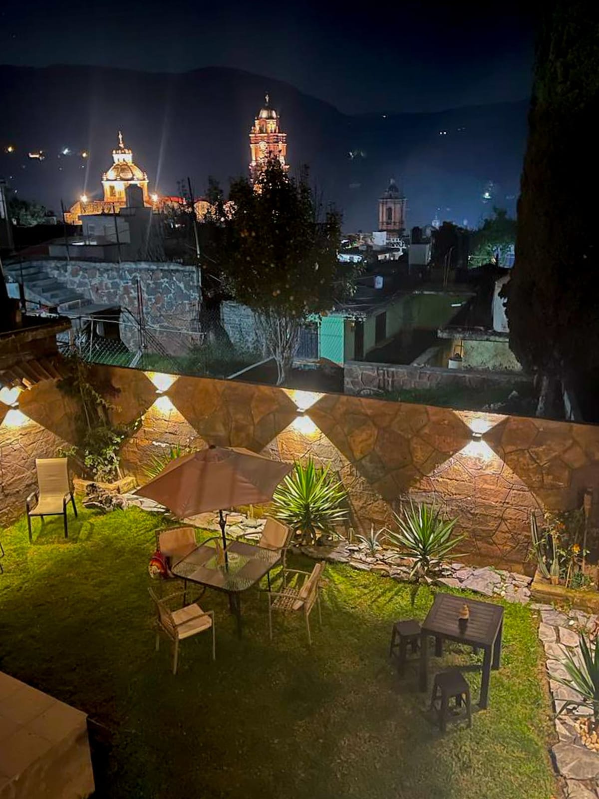 An outdoor garden area is illuminated at night, featuring a small table with four chairs. A large umbrella provides shade over the seating. The backdrop showcases a distant view of city lights and historical buildings, framed by gentle hills.