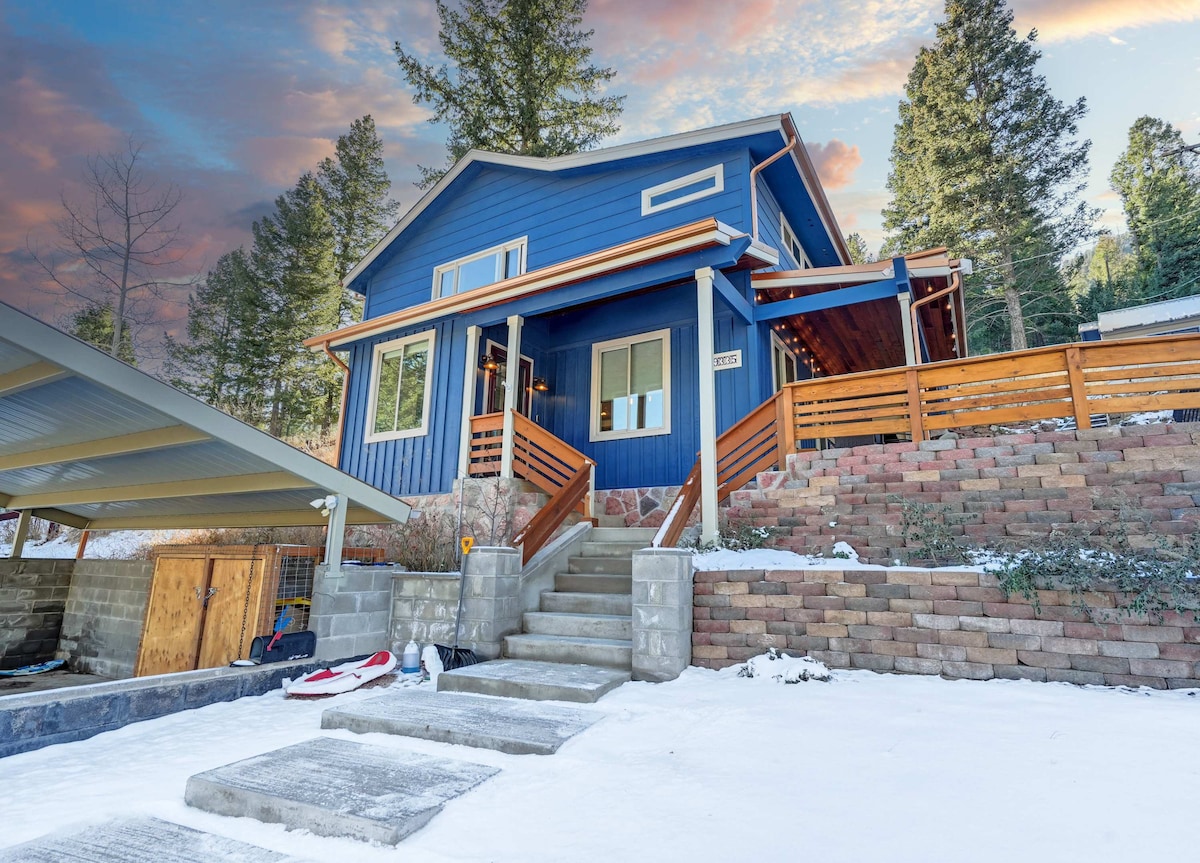 The exterior of the cabin showcases blue siding and a covered porch with wooden railings, nestled among snow-covered ground and evergreen trees. Concrete steps lead up to the entrance, which is framed by large windows, inviting natural light into the home.
