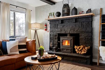 A cozy living area features a stone fireplace with a wood-burning stove at its center. A wooden coffee table with decorative items is positioned in front of a comfortable brown sofa adorned with various cushions. A soft lamp stands nearby, providing ambient lighting.