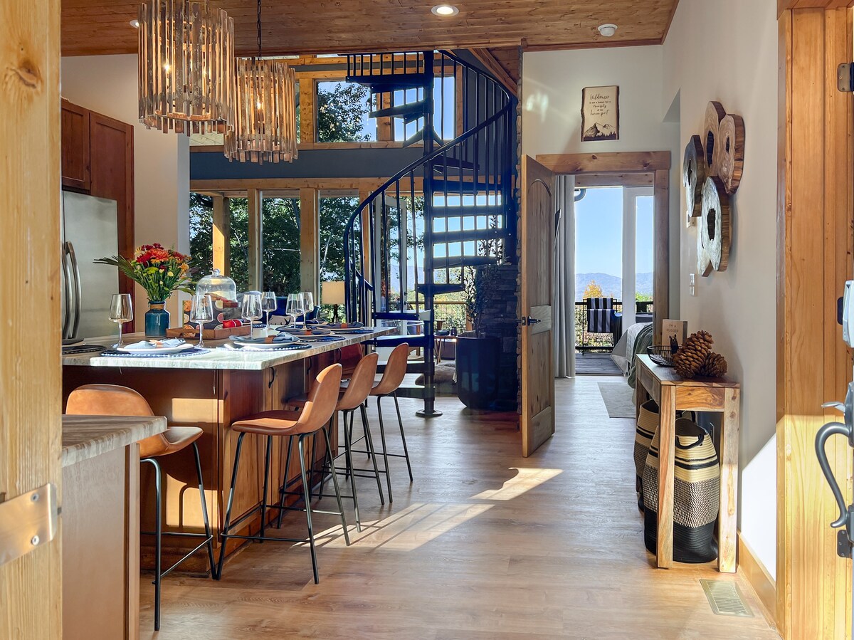 A modern kitchen with a large granite island featuring seating for seven. Pendant lights hang above, enhancing the natural light that floods the space. A spiral staircase leads to additional levels, while tall windows showcase beautiful mountain views beyond.