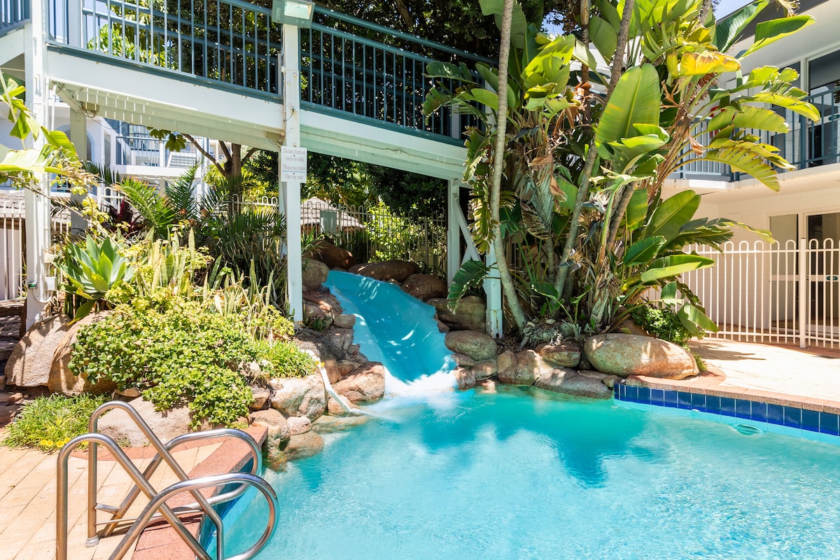 A resort-style pool is surrounded by tropical plants and a stone waterfall. A slide emerges from the upper level, emptying into the clear turquoise water. Nearby, a set of metal stairs offers access, inviting relaxation in this refreshing outdoor space.