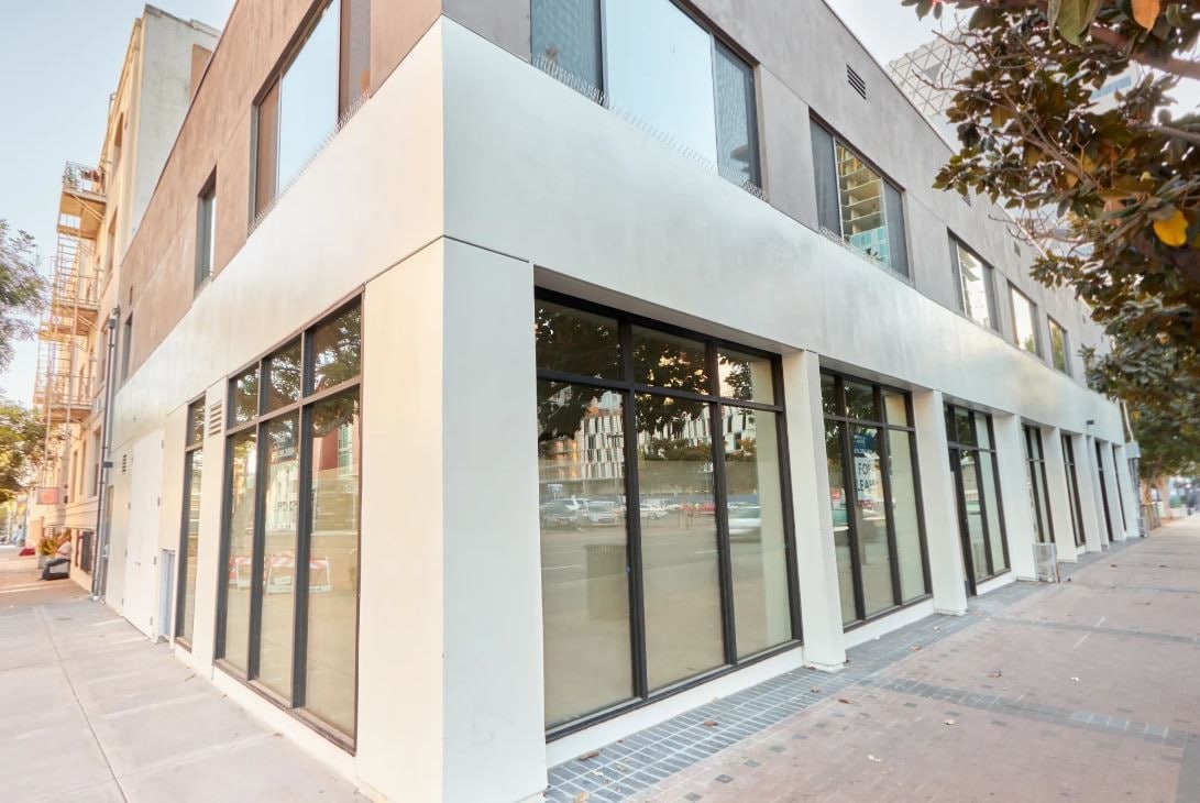 A two-story building showcases a modern exterior with large windows on the ground floor. The sleek design is complemented by a mix of light and dark materials, while the surrounding sidewalk contains patches of landscaping and urban elements.