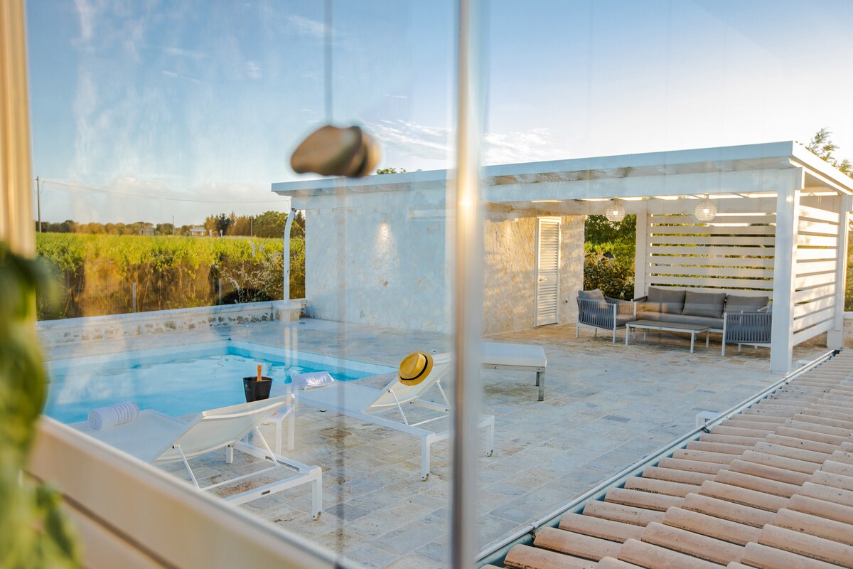 A clear view of the private pool area is presented, featuring several sun loungers arranged around the pool. A shaded seating area with modern furniture is visible in the background, creating a comfortable space for relaxation. The surrounding landscape is complemented by a tranquil sky.