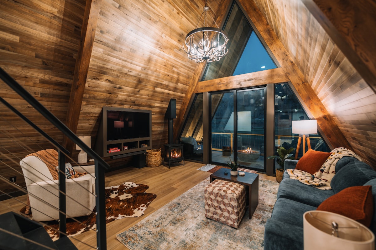 The living area features vaulted wooden ceilings and large windows that frame the view of the surroundings. A comfortable seating arrangement is highlighted, including a blue couch and an accent chair. A decorative coffee table sits on a patterned rug, and a cozy fireplace adds warmth to the space.