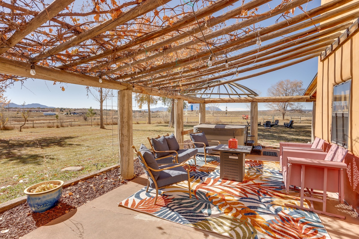 An outdoor patio area is seen, featuring a colorful rug beneath seating arrangements made up of various chairs and a coffee table. A pergola is visible overhead, adorned with climbing vines. Open space extends beyond, showcasing a view of the surrounding landscape.