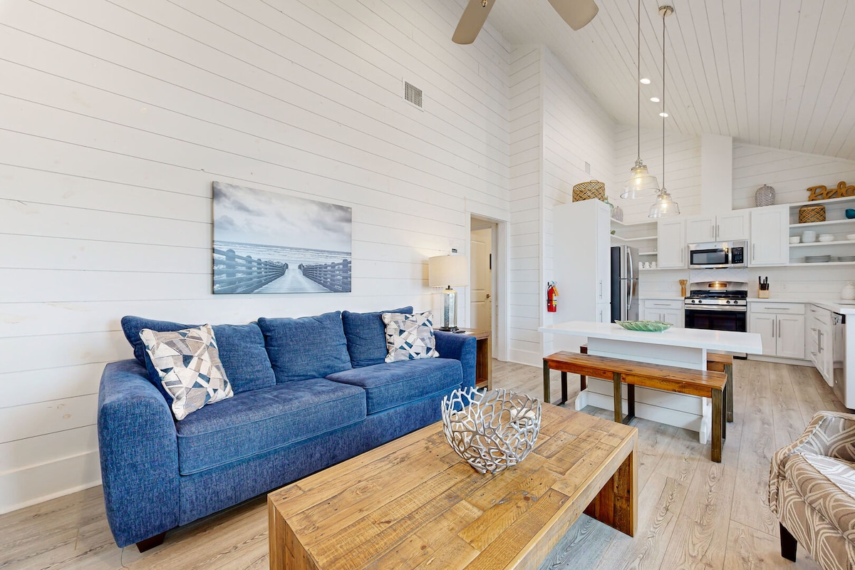 A spacious living area is featured, showcasing a large blue sofa with decorative pillows. A wooden coffee table occupies the center of the room, accompanied by an assortment of light-colored cabinetry and appliances in the kitchen area in the background.