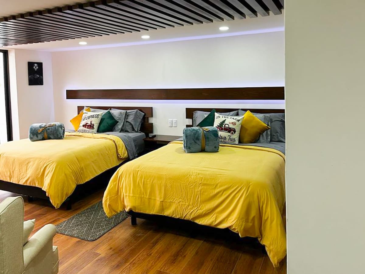 The spacious bedroom is furnished with two Queen Size beds, adorned with colorful throw pillows. Soft lighting illuminates the area, highlighting the wooden accents and floors. A comfortable seating area is positioned nearby for relaxation.