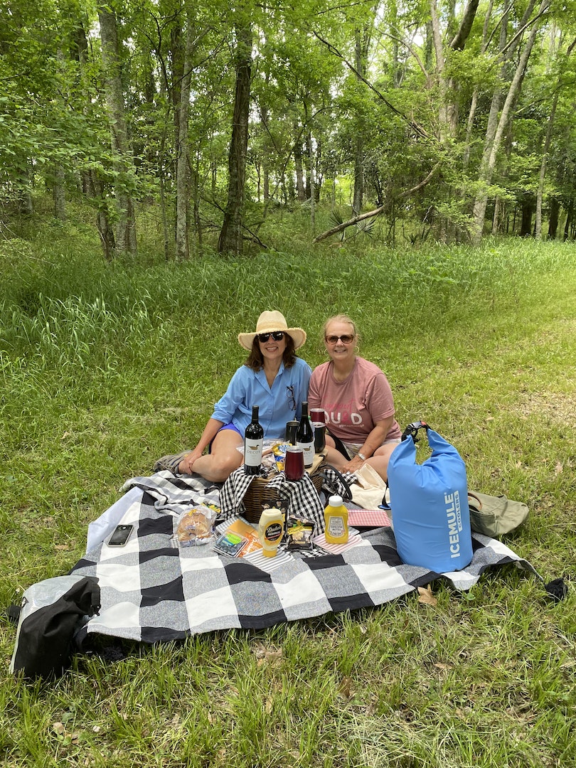 A picnic setup is arranged on a black-and-white checkered blanket nestled in a grassy area surrounded by lush greenery. Various food items and beverages are displayed on the blanket, enhancing a leisurely outdoor dining experience.