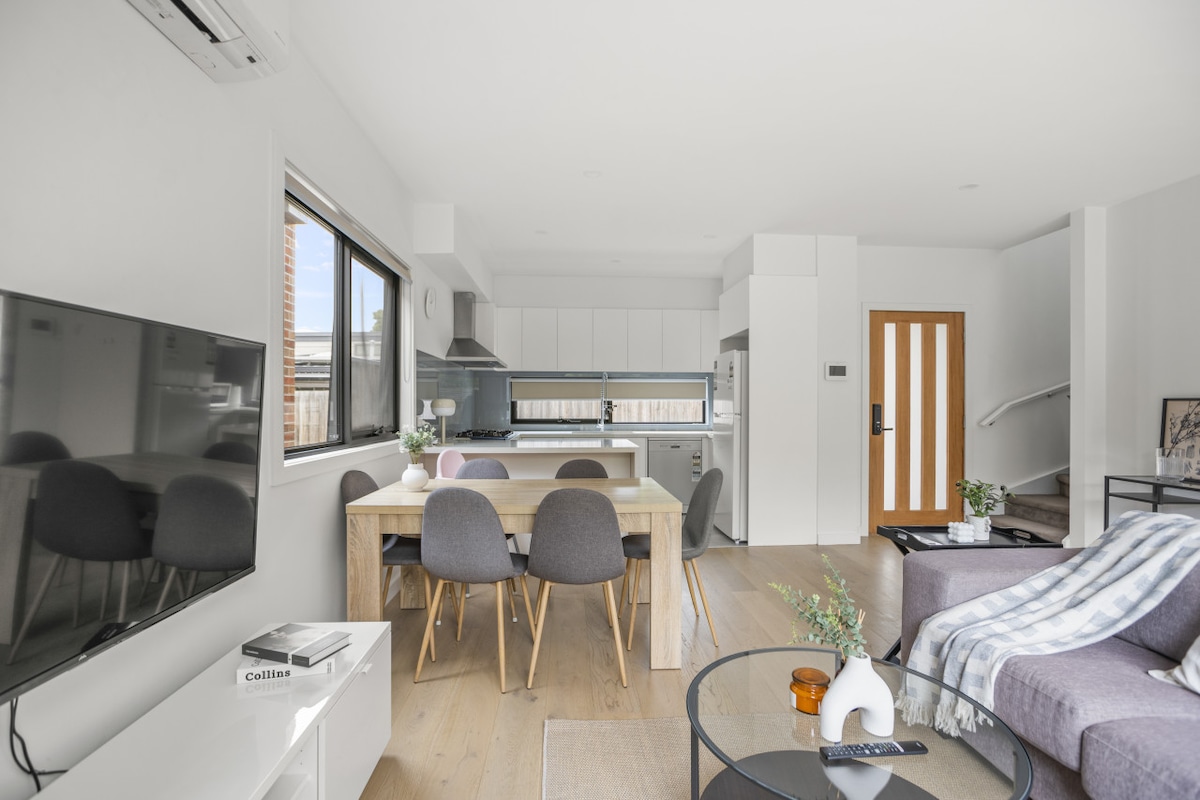 An open living space is presented, featuring a modern kitchen with sleek cabinetry and stainless steel appliances. A dining table with gray upholstered chairs is positioned in the center, while a comfortable seating area includes a plush sofa adorned with a soft throw. Natural light fills the room through large windows.