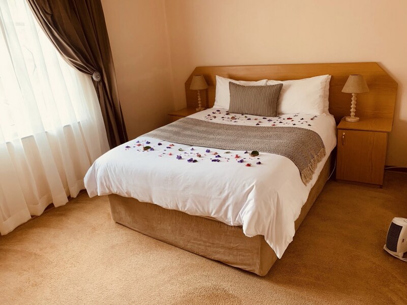 A cozy bedroom features a spacious bed adorned with decorative flower petals. Two bedside lamps provide gentle lighting. Soft beige carpeting complements the warm tones of the room, while sheer curtains allow natural light to softly filter in.