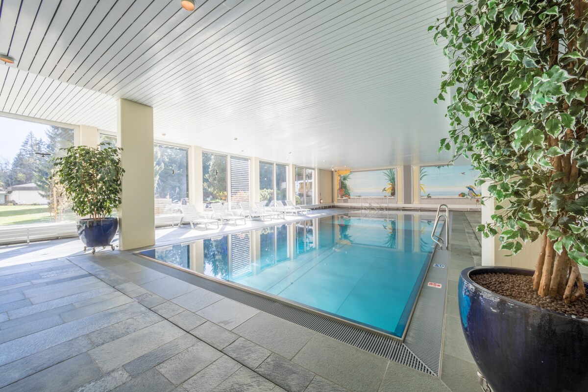 An indoor heated pool is situated within a spacious area, featuring large windows that allow natural light to illuminate the space. The pool is surrounded by comfortable lounge chairs, with decorative plants placed at the edges, creating a serene environment.