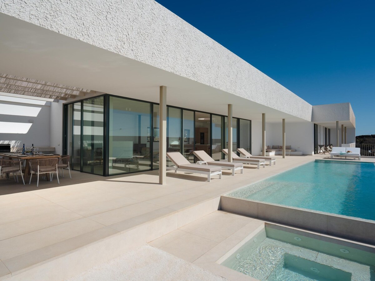 The modern villa features expansive glass walls that create a seamless connection between indoor and outdoor spaces. A heated pool with a jacuzzi is visible, surrounded by individual loungers placed on a spacious terrace. An inviting dining area is situated under a shaded structure.