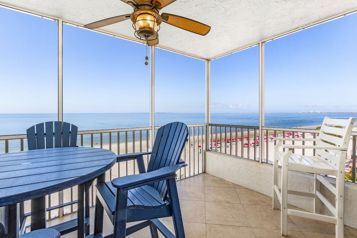 A screened-in balcony offers expansive views of the Gulf of Mexico. The outdoor seating area features a round table surrounded by blue adirondack chairs, while a white chair adds contrast. A ceiling fan hangs overhead, enhancing the coastal ambiance.