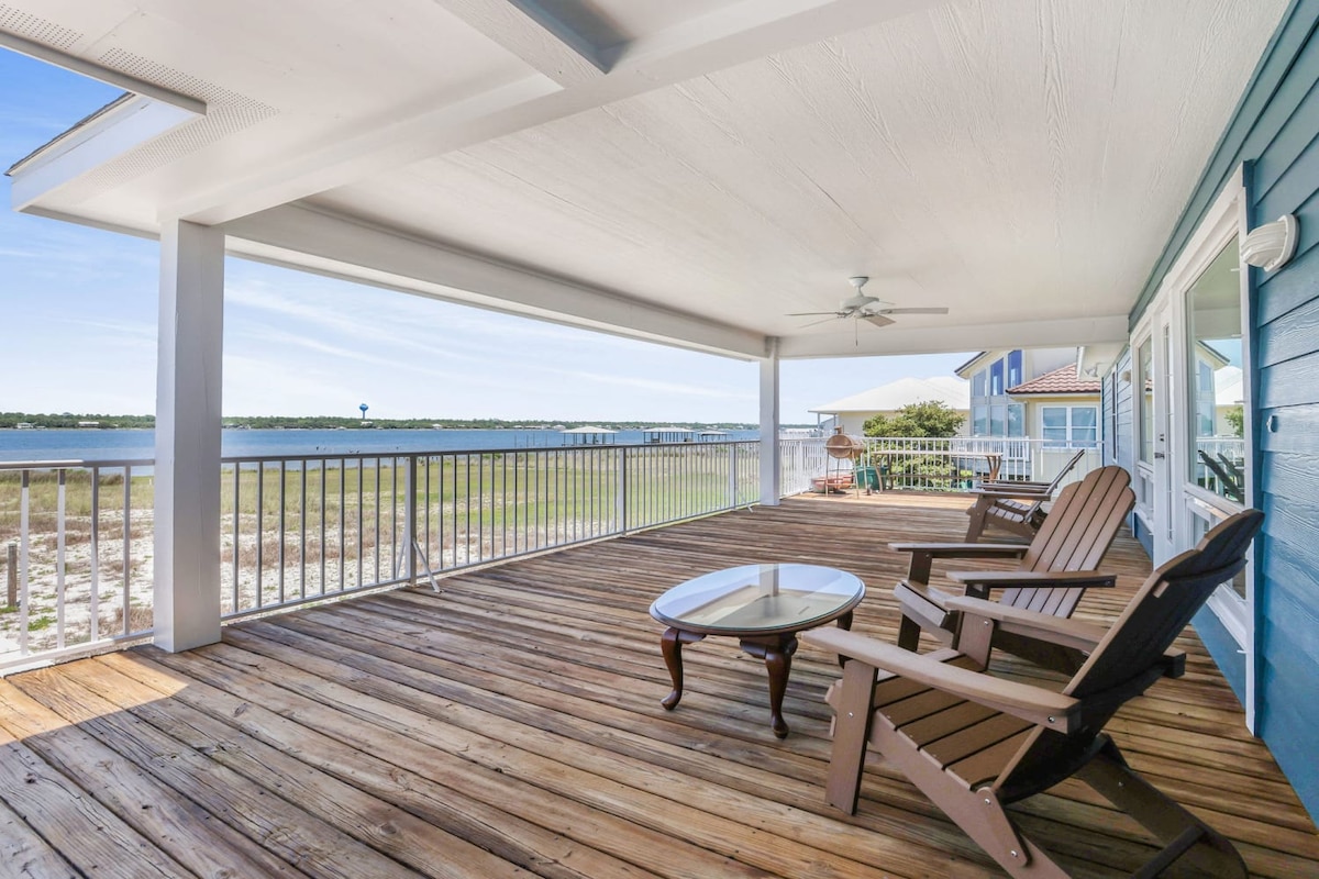 An expansive balcony features wooden flooring and a railing overlooking the lagoon. Two adirondack chairs are positioned near a small table, inviting relaxation while enjoying the water views. A ceiling fan hangs from the covered roof, providing additional comfort.