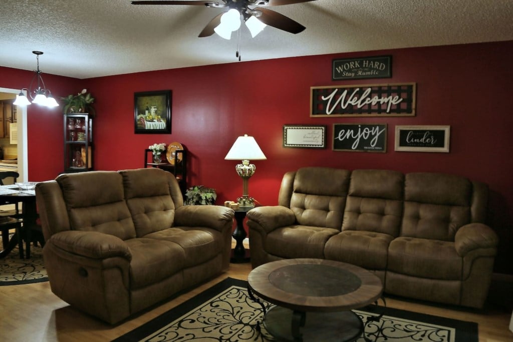 A cozy living room is highlighted by a warm red accent wall and comfortable seating. Two plush sofas face a circular wooden coffee table, while a standing lamp provides soft lighting. Wall decor includes welcoming signs, enhancing the inviting atmosphere.