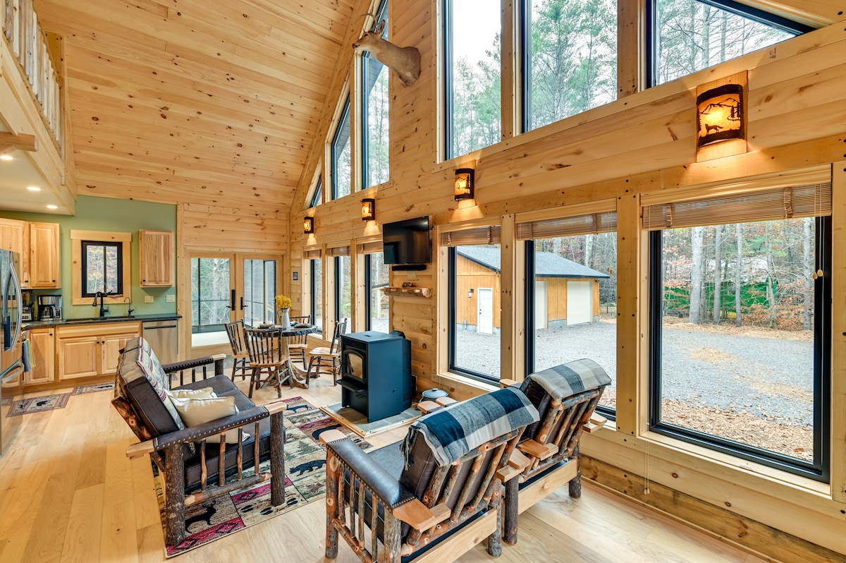 A spacious living area is surrounded by large windows, offering a view of the outdoor surroundings. Rustic wooden furniture includes comfortable seating and a coffee table. A modern wood-burning stove is positioned nearby, with soft lighting provided by wall sconces.