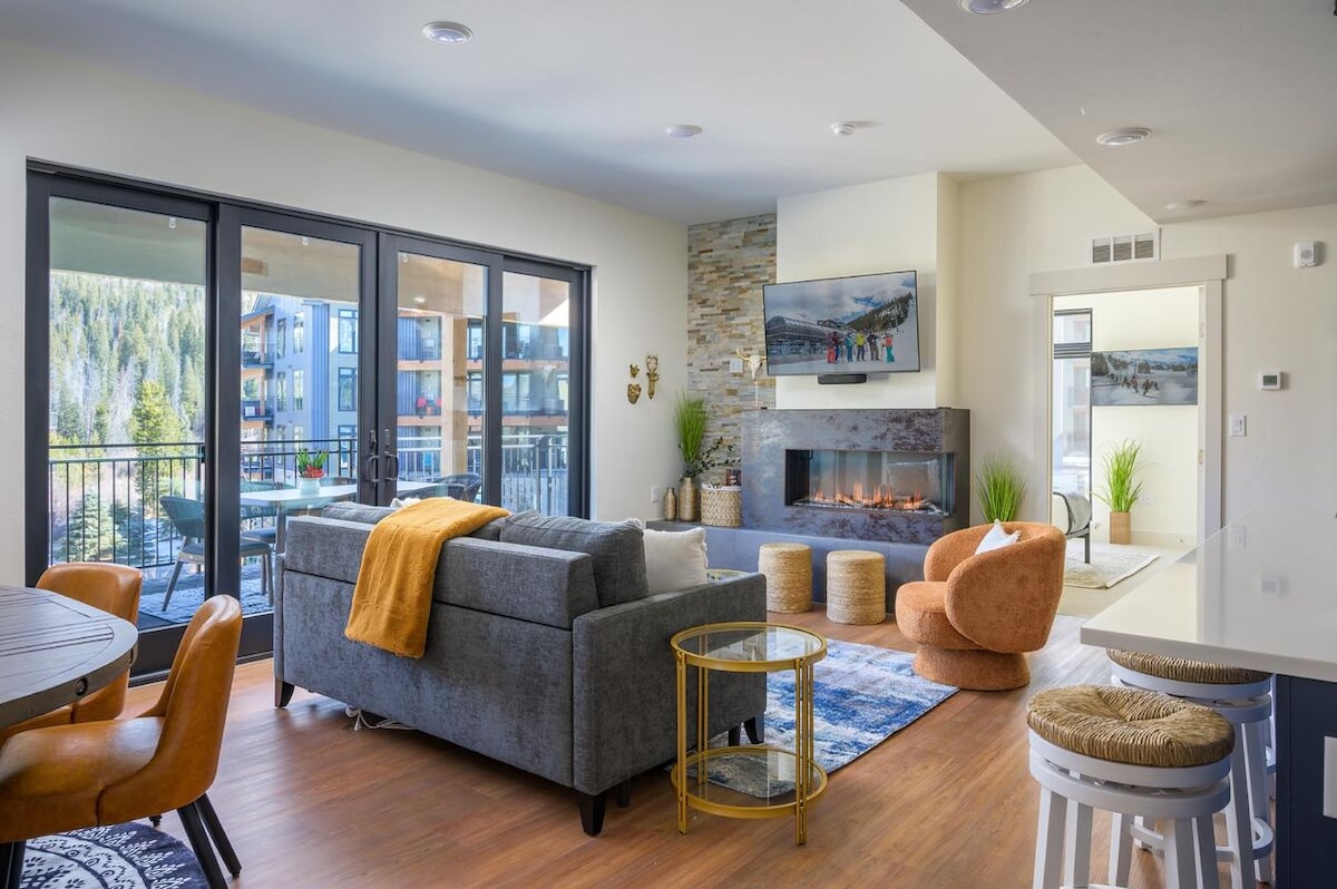 An open living space features a cozy sofa draped with a warm throw beside a sleek fireplace. Large glass sliding doors lead to a balcony, while a round dining table and a modern kitchen area can be seen in the background.