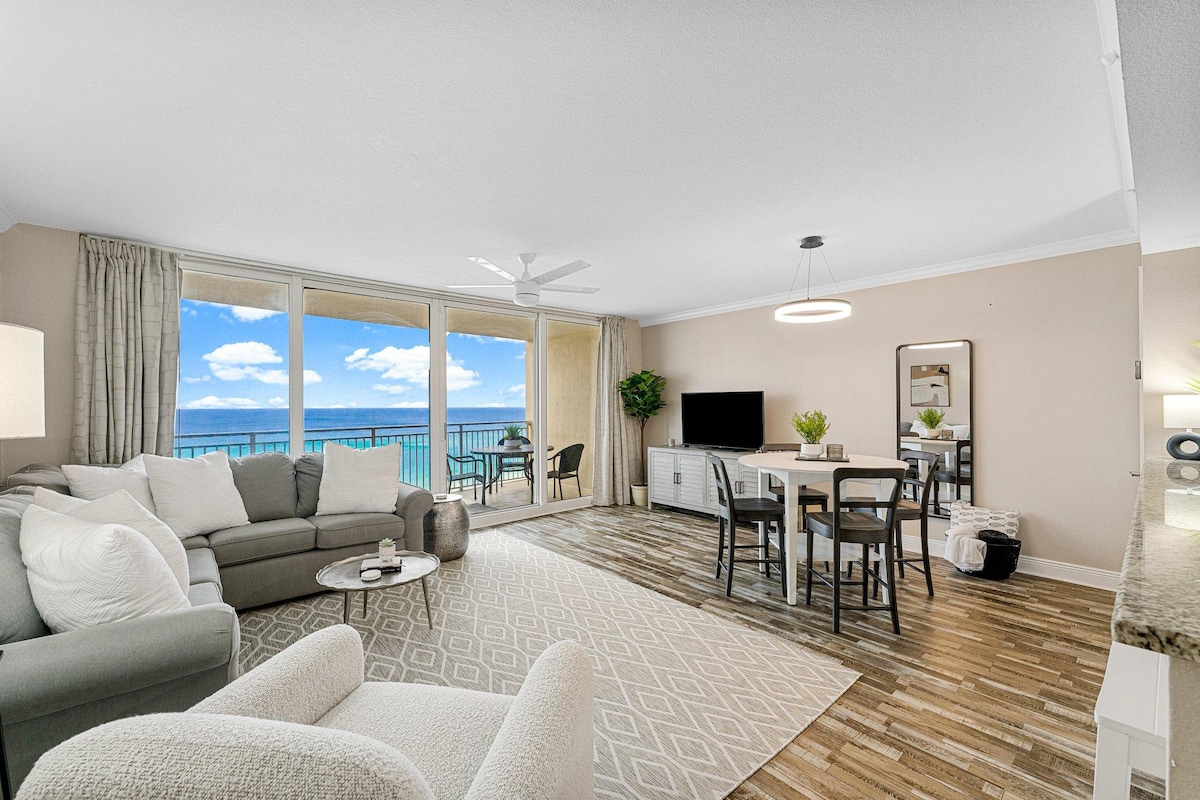 A spacious living area features large glass doors that provide magnificent views of the Gulf. Comfortable seating is arranged around a circular coffee table, with a dining area visible nearby. Light-colored walls and flooring enhance the bright, airy atmosphere created by natural light.