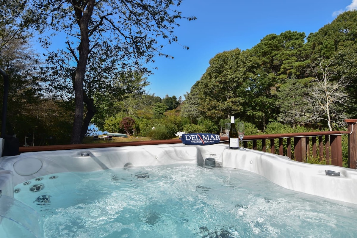 A four-person hot tub bubbles gently in a private backyard setting, surrounded by trees and open sky. A bottle of wine rests on the edge, adding to the tranquil ambiance of relaxation overlooking the lush scenery.