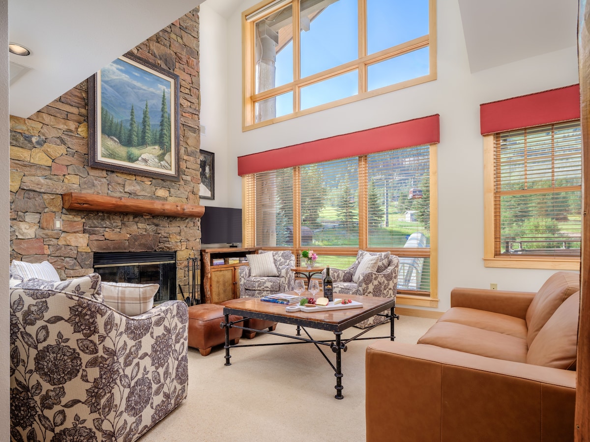 A well-furnished living area is highlighted by two windows allowing natural light to brighten the space. A stone fireplace adds a rustic touch, while plush seating is arranged around a coffee table. Art adorns the wall, enhancing the mountain retreat ambiance.